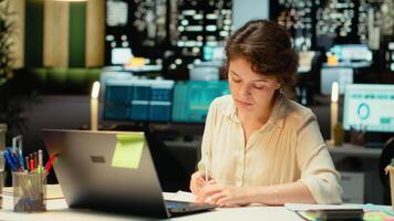 affärskvinna dokumentera grundläggande idéer från pc skärm på papper, planera och schemaläggning uppgifter sent på natt. anteckna med handstil för presentation den där speglar mål. kamera b. video