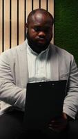 Vertical Black young man applicant getting called in for interview at HR office, sitting in the hallway and reviewing experience presentation on CV. Preparing for the hiring discussion. Camera A. video