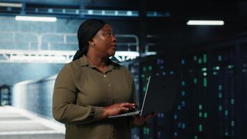 Admin walks between server farm rows, using laptop to upgrade rigs for increase in clients workload capacity. Data center worker configures hardware with notebook, improving processing speed, camera B video