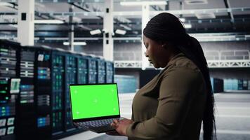 Server room admin uses green screen laptop to upgrade mainframes for increase in workload capacity. Data center worker configures gear with mockup notebook, improving processing speed, camera A video