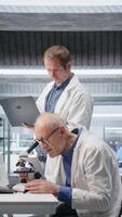 verticale vecchio uomo scienziato utilizzando microscopio con un' collega per clinico studia. scorrevole il campione vassoio per indagine e supporto genetica ricerca, Avanzate analisi per assistenza sanitaria. telecamera un. video