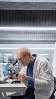Vertical Male researcher at microscope analyzes bioscience samples from lab tray. Focus on cellular observation and detailed molecular study emphasizes the scientific method, biochemistry and data analysis. Camera A. video