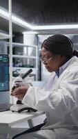 vertical africano americano mulher analisando molecular amostras dentro uma bio laboratório, usando profissional microscópio. cuidados de saúde e bioquímica experimentos representar científico método e descoberta. Câmera uma. video