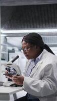 vertikal forskare analyserar prover i laboratorium miljö med mikroskop och kemikalier. kvinna expert- framåt genetik forskning, kombinerande laboratorium analys med biokemi studier. kamera a. video