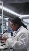 Vertical Black female expert analyzing samples in chemistry lab with microscope and observation tools. Scientist advancing genetics research, pathology analysis with biochemistry studies. Camera A. video