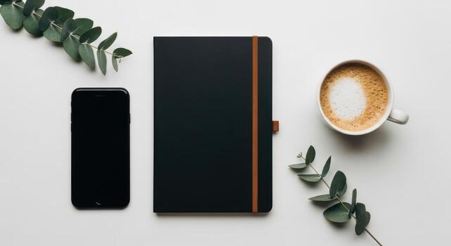Flat lay of a black notebook, smartphone, coffee cup, and eucalyptus leaves on a white surface photo
