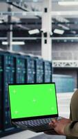 Vertical Server room admin uses green screen laptop to upgrade mainframes for increase in workload capacity. Data center worker configures gear with mockup notebook, improving processing speed, camera A video