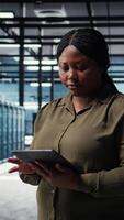 Vertical Data center software developer monitors data center metrics on tablet to enhance operational effectiveness. Woman in server room using app device analytics to predict performance, camera A video