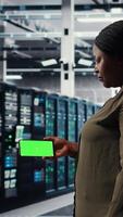 Vertical Server hub software developer uses green screen smartphone to watch tutorial on how to upgrade rig. African american woman in data center follows guide on mockup phone to improve speed, camera A video