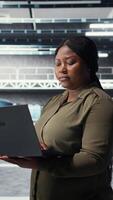 Vertical Technician in server room using laptop to upgrade rigs for increase in clients workload capacity. African american woman in data center configures equipment, improving processing speed, camera A video