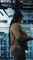 Vertical Engineer walks in server hub, using laptop to upgrade rigs for increase in clients workload capacity. Data center employee configures equipment with notebook, improving processing speed, camera A video