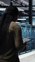 Vertical High tech data center technician uses tablet to develop disaster recovery plans, minimizing downtime. IT employee walking in server room, enhancing data protection protocols using device, camera A video
