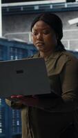 Vertical Smiling engineer walking in data center with laptop, checking hardware for system stability. Upbeat employee overseeing server farm with notebook, configuring infrastructure, camera B video