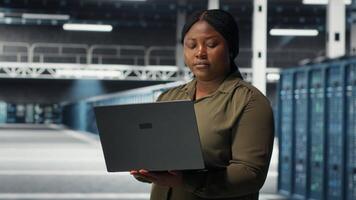 Technician in server room using laptop to upgrade rigs for increase in clients workload capacity. African american woman in data center configures equipment, improving processing speed, camera B video