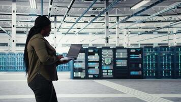 dati centro lavoratore utilizzando il computer portatile per gestire Software aggiornamenti per sistema stabilità. africano americano donna nel server camera configurazione infrastruttura per ottimale prestazione, telecamera un' video
