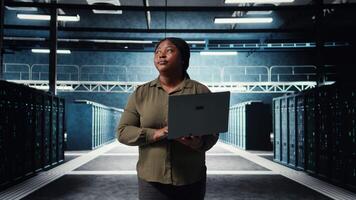 Admin walks between server farm rows, using laptop to upgrade rigs for increase in clients workload capacity. Data center worker configures hardware with notebook, improving processing speed, camera A video