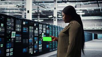 Server hub software developer uses green screen smartphone to watch tutorial on how to upgrade rig. African american woman in data center follows guide on mockup phone to improve speed, camera A video