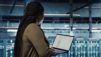 Engineer walks in server hub, using laptop to upgrade rigs for increase in clients workload capacity. Data center employee configures equipment with notebook, improving processing speed, camera B video