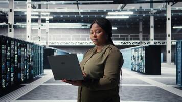 Technician in server room using laptop to upgrade rigs for increase in clients workload capacity. African american woman in data center configures equipment, improving processing speed, camera A video