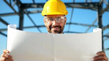 Lycklig konstruktion arbetstagare på en solig dag på en byggnad webbplats innehav ritningar med strukturell stål ramverk i de bakgrund video