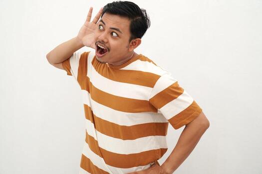 Young Asian man in striped tshirt posing with a curious expression, holding one hand behind his ear as if trying to listen closely, standing against a white background.., photo