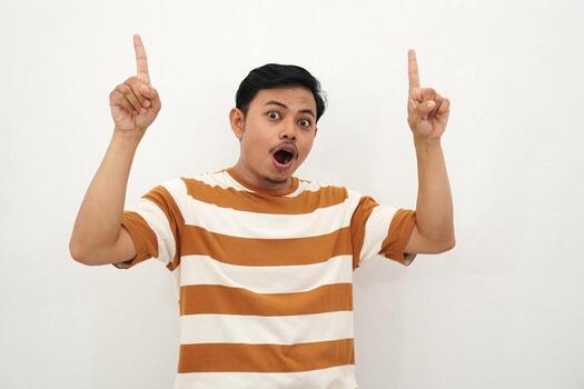 Young Asian man in white and brown striped t-shirt smiling and pointing up with both index fingers enthusiastically, posing with a cheerful and eye-catching gesture against a white background, photo