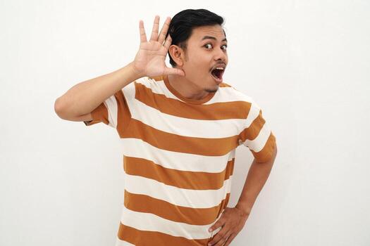 Young Asian man in striped tshirt posing with a curious expression, holding one hand behind his ear as if trying to listen closely, standing against a white background., photo