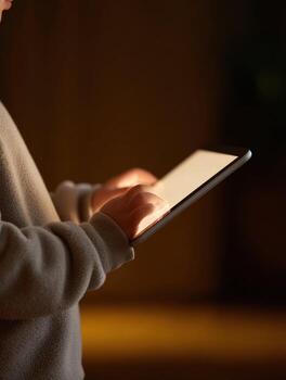 Focused child hand holding tablet in dark. screen light illuminates youth, deeply engaged with modern technology indoors, creating calm atmosphere photo