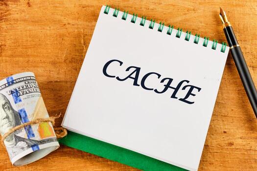 A notebook with the word CACHE written prominently is placed on a wooden table next to rolled currency and a pen, highlighting the importance of cache in modern finance photo