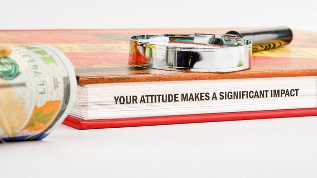 A motivational book rests on a surface, adorned with a magnifying glass alongside a rolled banknote. The text highlights the importance of attitude in achieving success and fulfillment photo