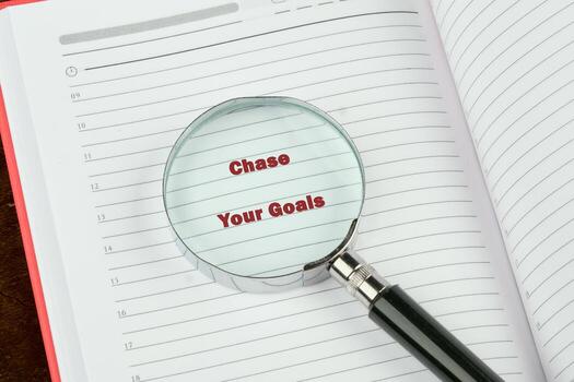 A magnifying glass rests upon an open notebook its lens highlights the text Chase Your Goals printed on the lined pages. It suggests planning and ambition photo