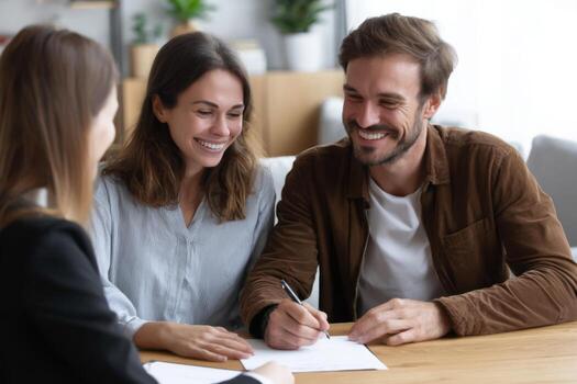 A couple sits at a wooden table in a bright office, smiling while reviewing paperwork with a consultant. They appear excited and engaged in the conversation, planning their next steps photo