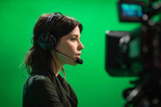 A broadcaster is focused on her work in a studio with a green backdrop. She wears a headset and microphone while operating a camera. The atmosphere is professional and concentrated photo