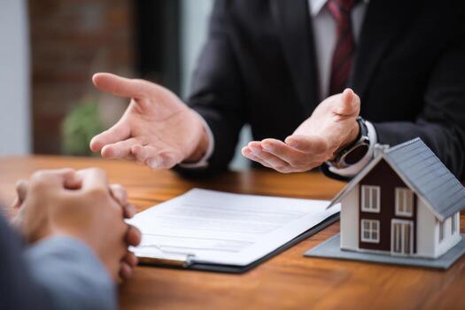 A professional real estate agent presents property information to a client, engaging in discussion at a desk with a small house model and documents in an office environment photo