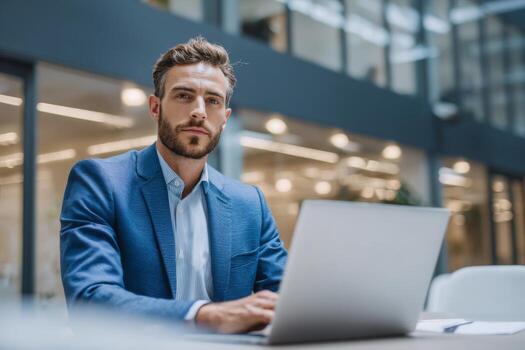 A man in business attire is seated at a sleek desk, diligently working on his laptop in a contemporary office. Natural light streams through large windows, enhancing the productive atmosphere photo