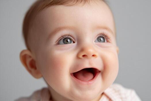 A cheerful baby with sparkling blue eyes smiles widely, showcasing pure joy. The soft neutral background enhances the babys delightful expression during a playful moment photo