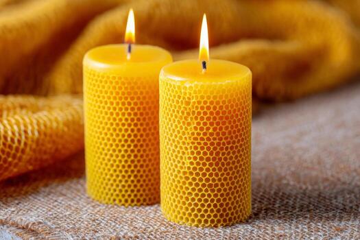 Two vibrant yellow beeswax candles are lit and placed on a textured surface, showcasing their honeycomb pattern. The background features a soft, warm fabric adding to the cozy ambiance photo