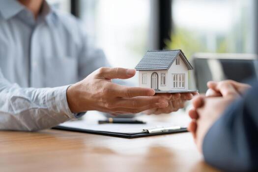 Two hands are seen exchanging a small house model during a real estate discussion in a bright office setting. The atmosphere feels professional and focused on property transactions photo