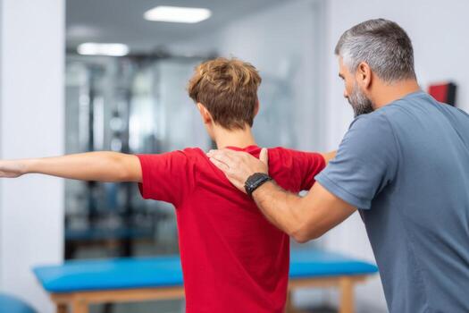 A personal trainer guides a young boy in a gym, focusing on body posture and exercise technique. The trainer demonstrates and offers support. Bright lighting enhances the training environment photo