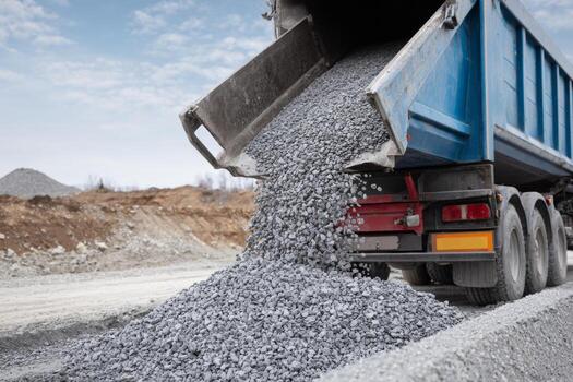 A dump truck is unloading a large quantity of gravel at a construction site during daylight. Heavy machinery and earthworks are visible in the background photo