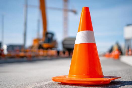 Bright orange traffic cone is positioned on the ground to signal construction activity. Heavy machinery and workers can be seen in the background, indicating ongoing work photo