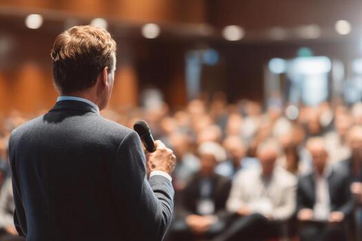 A speaker stands at the forefront, holding a microphone and addressing a large audience during a business conference. Attendees are focused and attentive in a well-lit hall photo