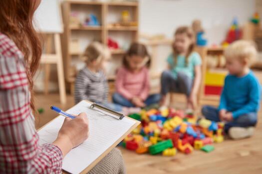 In a bright classroom, a teacher takes notes while four children play with colorful building blocks on the floor, promoting creativity and teamwork among them photo