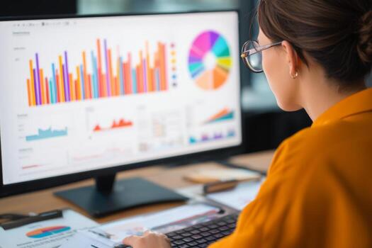 A professional woman wearing glasses studies vibrant data charts on a computer screen in a modern office setting during the day. Papers and a keyboard are on the desk photo