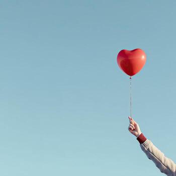 Hand holds a red heart-shaped balloon against a clear blue sky. A whimsical image photo