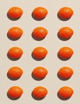 Fifteen orange, pebble-like objects are arranged in a grid, casting small shadows on a tan surface photo