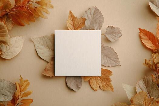 Square blank card surrounded by autumn-colored leaves, arranged symmetrically on a tan surface photo