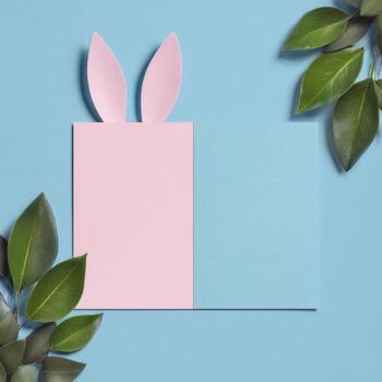 Pink paper rabbit ears, a card split by color, adorned by green leaves on blue photo
