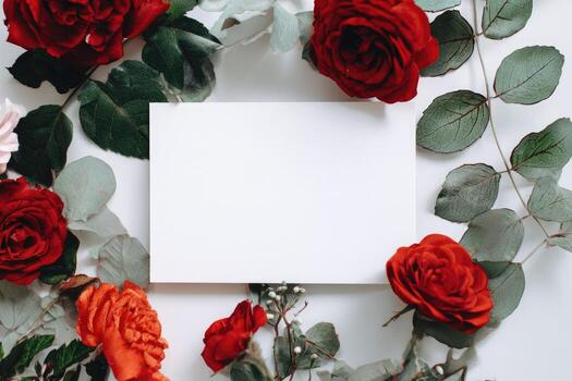 Overhead view of white blank card surrounded by red roses, green leaves, and a white backdrop photo
