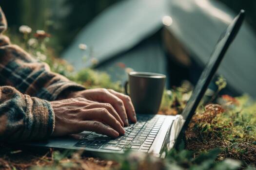 Close-up of hands typing on a laptop, sitting on the grass near a tent in a forest. Man is working outdoors using a computer. Close-up of a man using a notebook and drinking coffee, camping in nature. photo